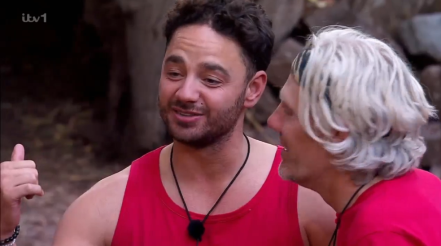 Two men in red tank tops talking, one with dark hair and a beard, the other with white hair.