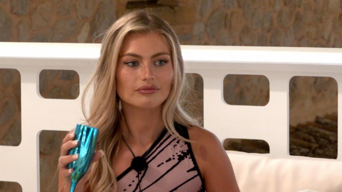 A woman with long blonde hair holds a blue champagne flute in a scene from Love Island.