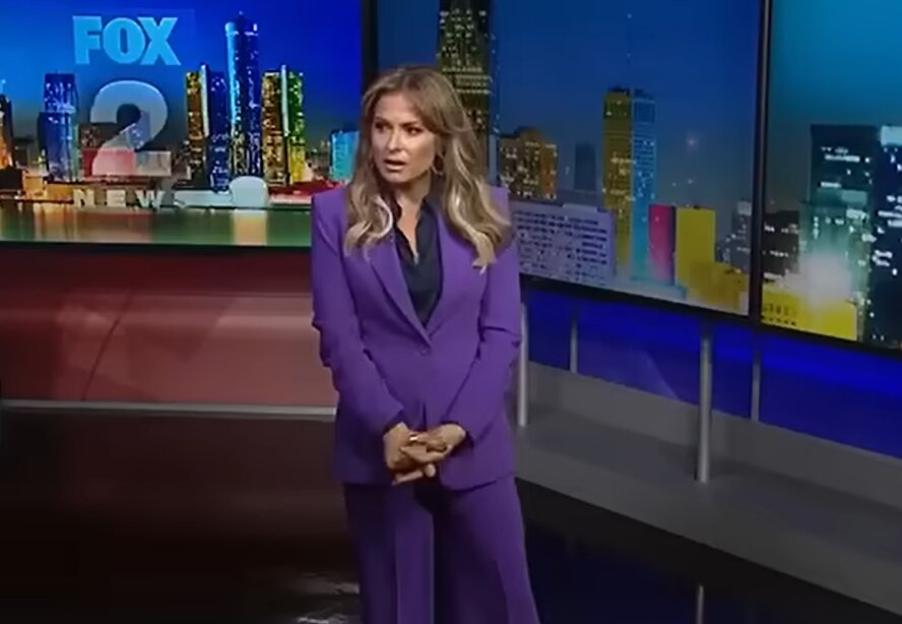 A female news anchor in a purple suit stands in front of a news desk with a city skyline backdrop.