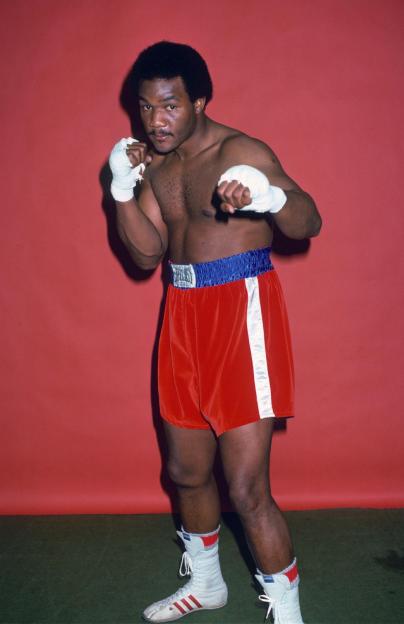 Heavyweight boxer George Foreman in a fighting stance with bandaged hands, red shorts, and white boxing boots.