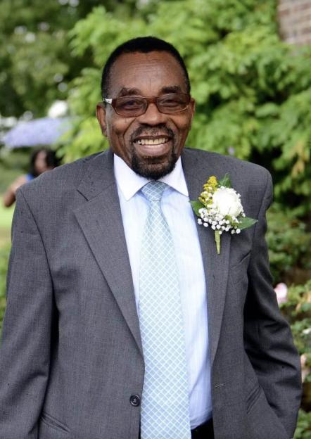 Franklin Agbotui, a smiling man with a short beard, glasses, light blue shirt, patterned blue tie, grey suit jacket, and white boutonnière.