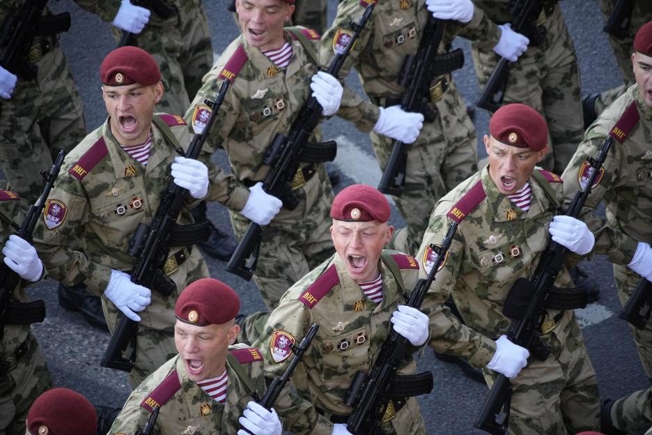 A group of soldiers in camouflage, maroon berets, and white gloves march with rifles.