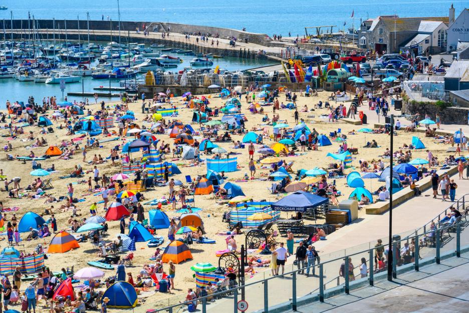 A crowded beach with sunbathers and families under colorful umbrellas and tents next to a marina filled with boats.