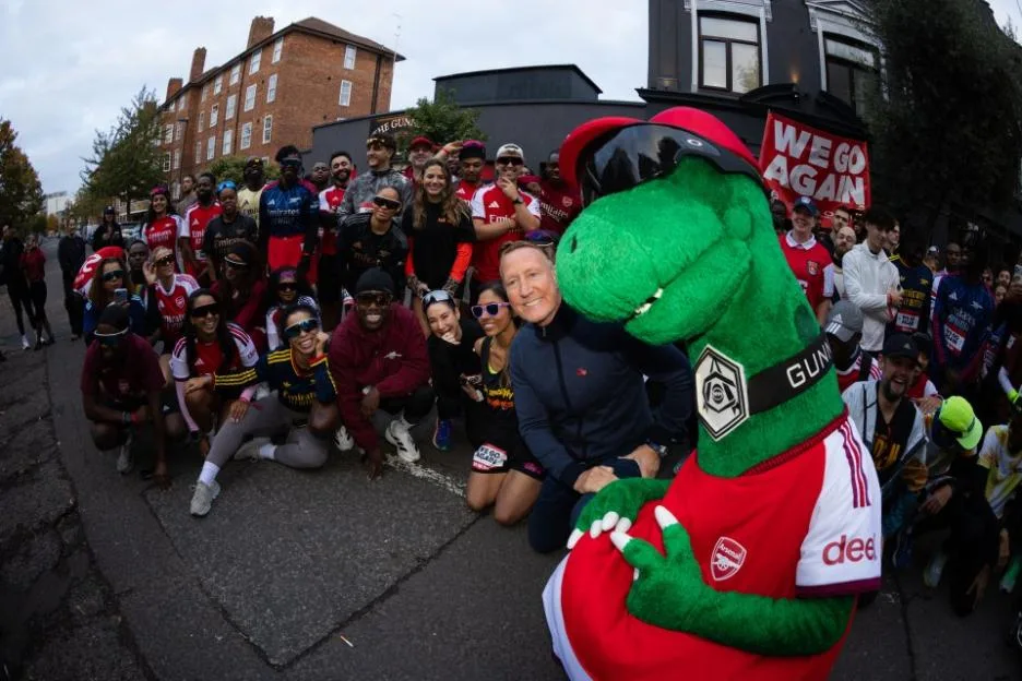 Arsenal fans and a green dinosaur mascot posing for a group photo on a street.