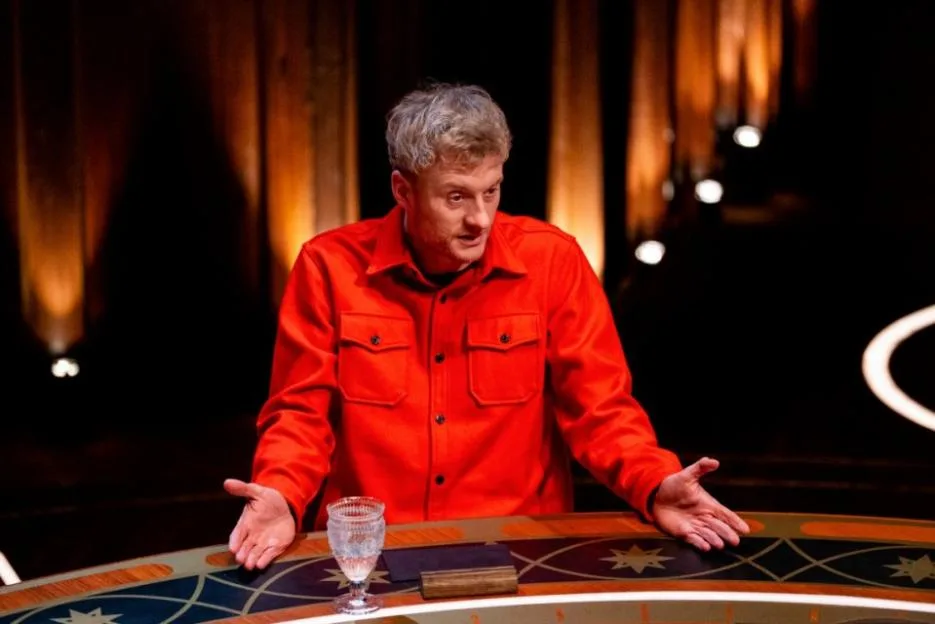 A man wearing a red shirt sitting at a round table with his hands outstretched.