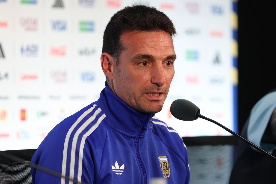 A man in a blue track jacket with white stripes and an Argentina football patch speaks into a microphone at a press conference.