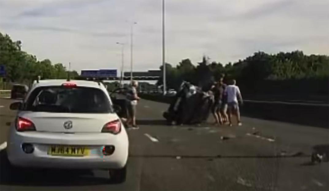 Several people stand around a dark, flipped-over car on a motorway with debris scattered around it.