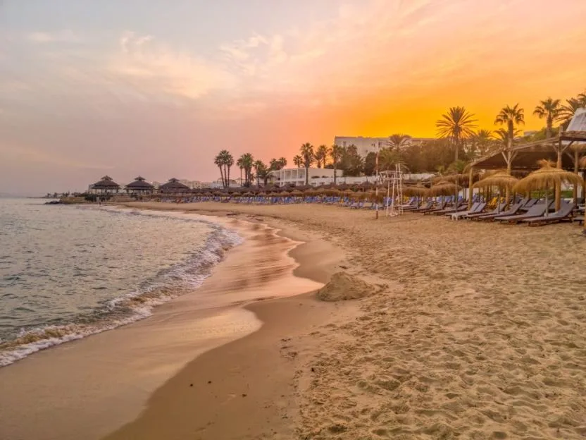 Landscape of a sandy beach in Hammamet, Tunisia at sunset, featuring thatched umbrellas, sun loungers, and palm trees along the coast.