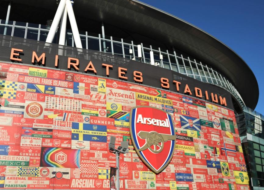 The exterior of Emirates Stadium with the word "EMIRATES STADIUM" on top and a large Arsenal logo and banners from various Arsenal fan clubs around the world below.