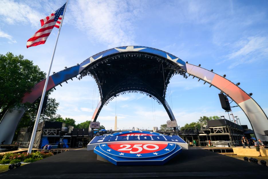 An American flag flies above a stage with "FREEDOM 250" on it, with the Washington Monument in the background.