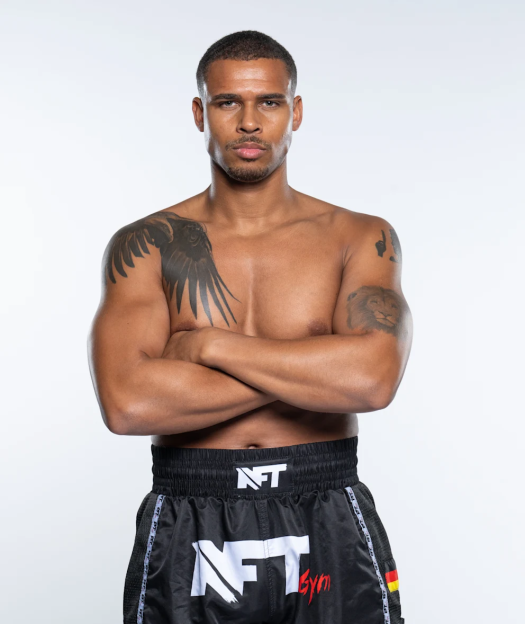 A shirtless man with tattoos on his arms, wearing black shorts with "NFT Gym" written on them, stands with his arms crossed.