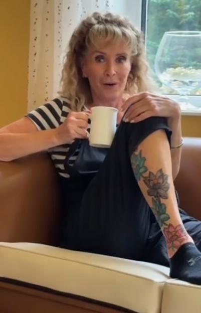 Beverley Callard sitting on a couch with a coffee mug, wearing a striped shirt and overalls, revealing lotus flower tattoos on her leg.