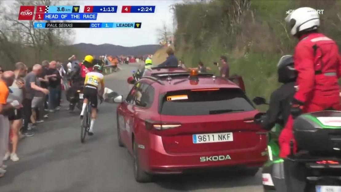 A cyclist with bib number 61, Paul Seixas, wearing a yellow and white jersey, riding ahead of a red Skoda car during a race, with spectators on the side of the road.