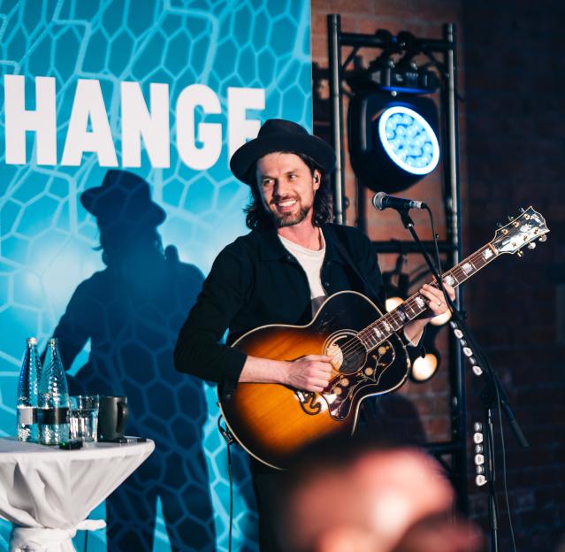 A smiling male musician with a hat playing an acoustic guitar on stage, with a turquoise backdrop.