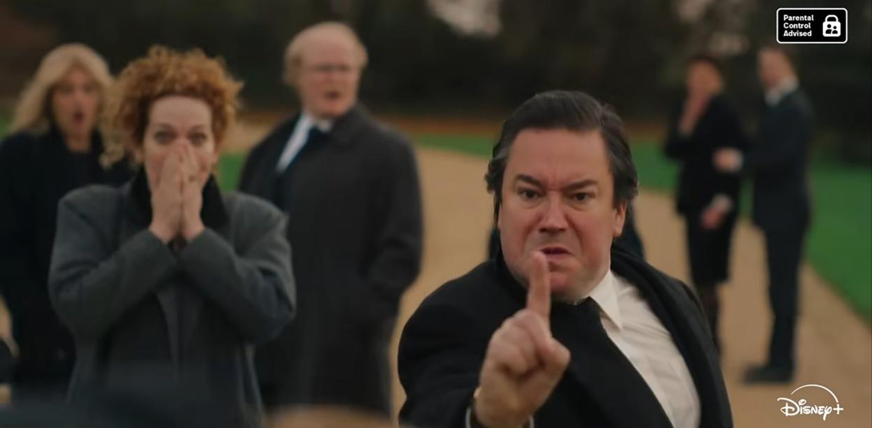A man in a suit jacket and white shirt pointing his finger in frustration with several other people looking surprised or concerned in the background.