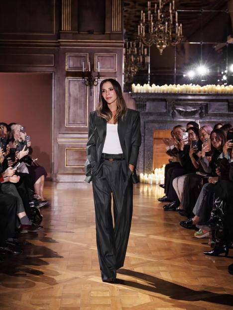 Victoria Beckham walking the runway at a fashion show, wearing a dark gray pantsuit with a white undershirt.