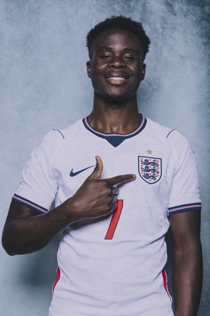 Bukayo Saka smiling and pointing to the England Football Association logo on his jersey.
