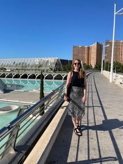 A woman in a black top and floral skirt stands on a bridge with modern architecture and pools in the background.