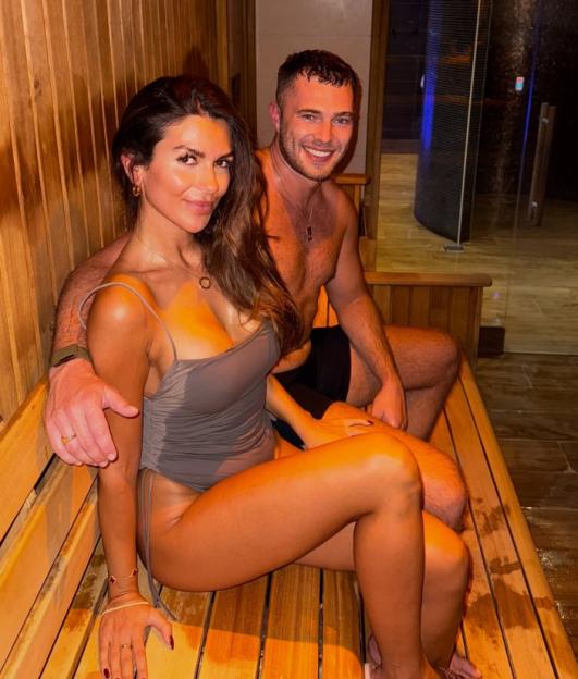 A smiling couple sits on a wooden bench, appearing to be in a sauna or spa.