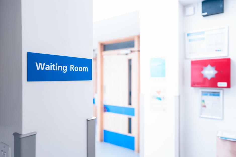 A blue "Waiting Room" sign on a white wall in a GP surgery with blurred doctor's offices in the background.