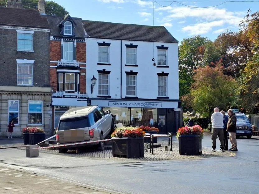 A silver Range Rover has crashed into a paved area, knocking down the Market Cross in front of a shop named Mahoney Hair Company.