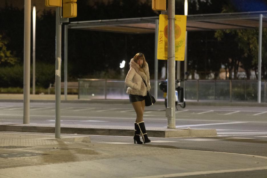 A woman in a fur-trimmed jacket, short skirt, and boots stands on a street corner at night in Barcelona, Spain.