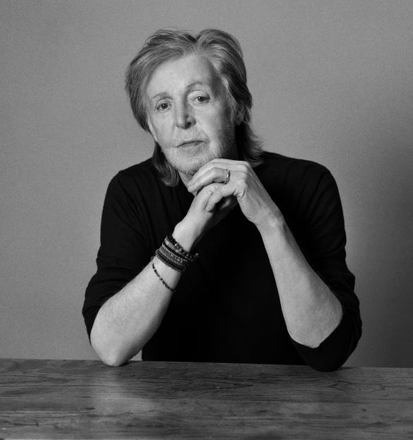 Black and white portrait of Paul McCartney looking directly at the camera with hands clasped under his chin.