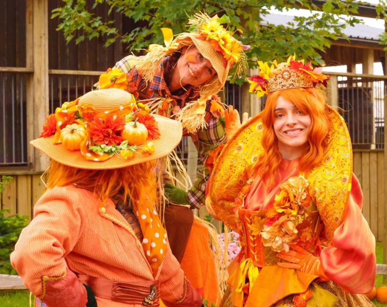 Three people in festive autumn costumes, with one dressed as a scarecrow and another wearing a crown and orange costume.