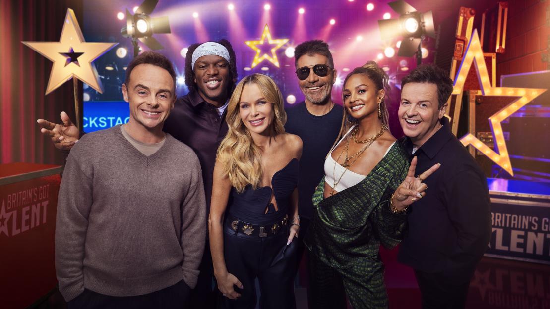 Simon Cowell wearing sunglasses, a dark blue t-shirt, and dark pants, smiling on the Britain's Got Talent set.