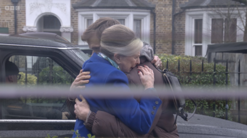 An older woman in a blue coat hugging a younger person in a brown coat with a black handbag, as a third person is seen in the background through a car window.