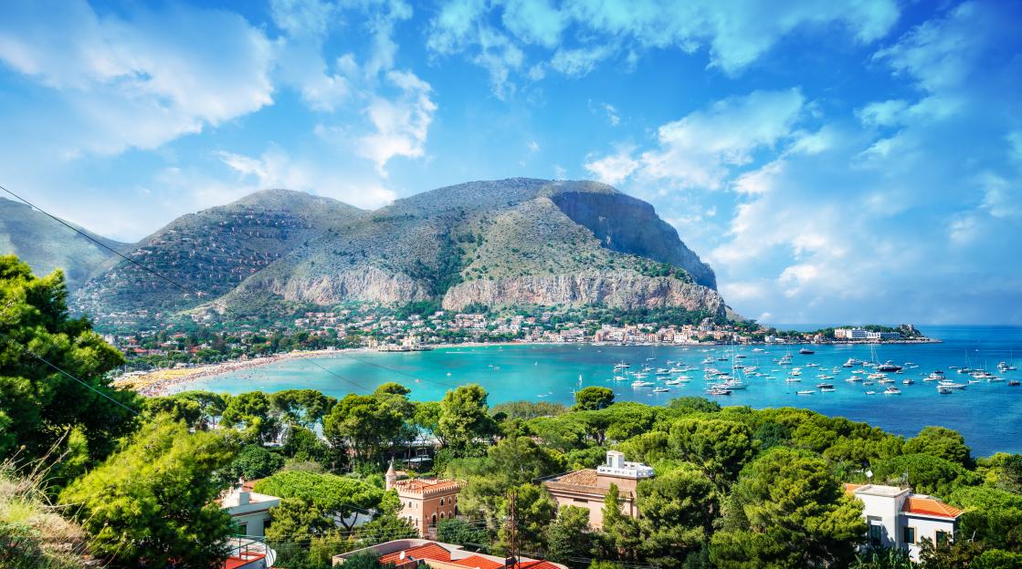 View of the Gulf of Mondello and Monte Pellegrino in Palermo, Sicily, Italy, showing the beach, boats in the water, and houses nestled in the mountains.