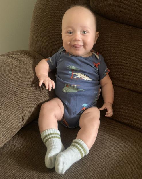 A baby wearing a dinosaur-print onesie and striped socks sits on a brown couch.