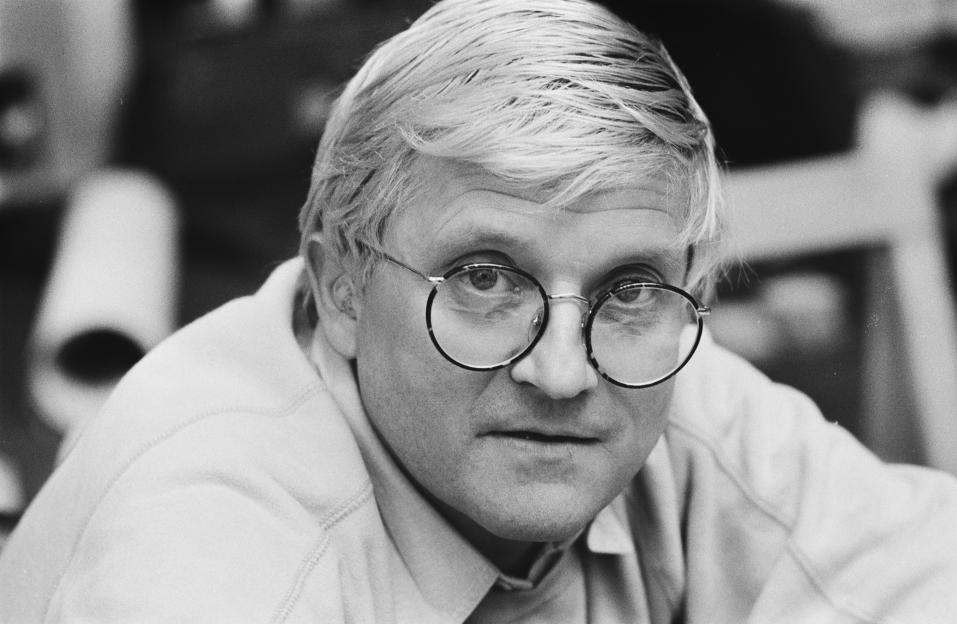 A black and white portrait of artist David Hockney wearing round glasses.