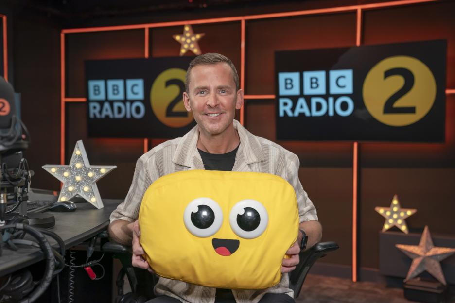 Scott Mills holding a yellow pillow with large eyes and a smiling mouth, promoting BBC Radio 2's takeover of CBeebies Bedtime Stories.