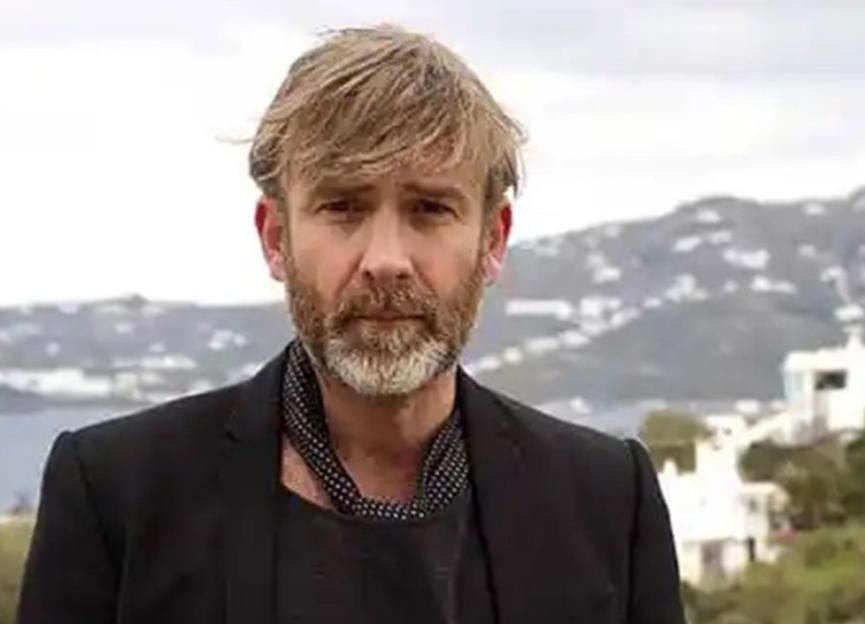 Dean Aslett, wearing a black blazer and t-shirt, against a hillside village background.