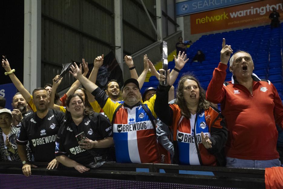 Fans at a London Broncos game with arms raised, cheering.