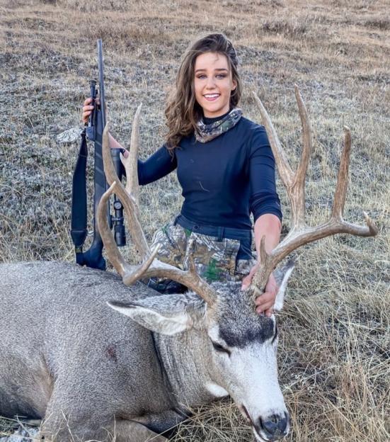 A young woman with a rifle poses with a large dead deer.