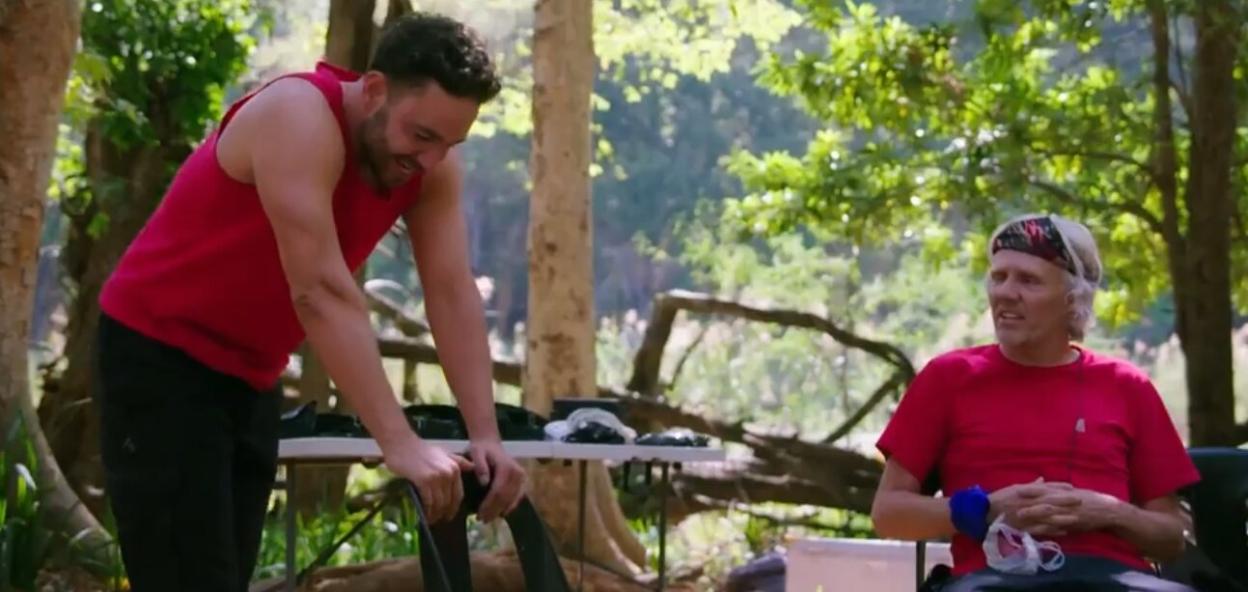 Adam Thomas in a red top standing in front of an older man in a red shirt with a bandana in a wooded area.
