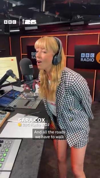 Sara Cox in a checkered jacket and headphones, singing into a microphone in a BBC Radio studio.