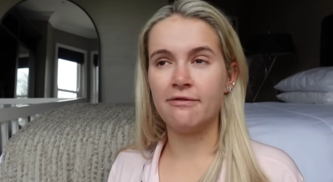 Molly-Mae Hague in a light pink top, speaking while sitting on a bed.