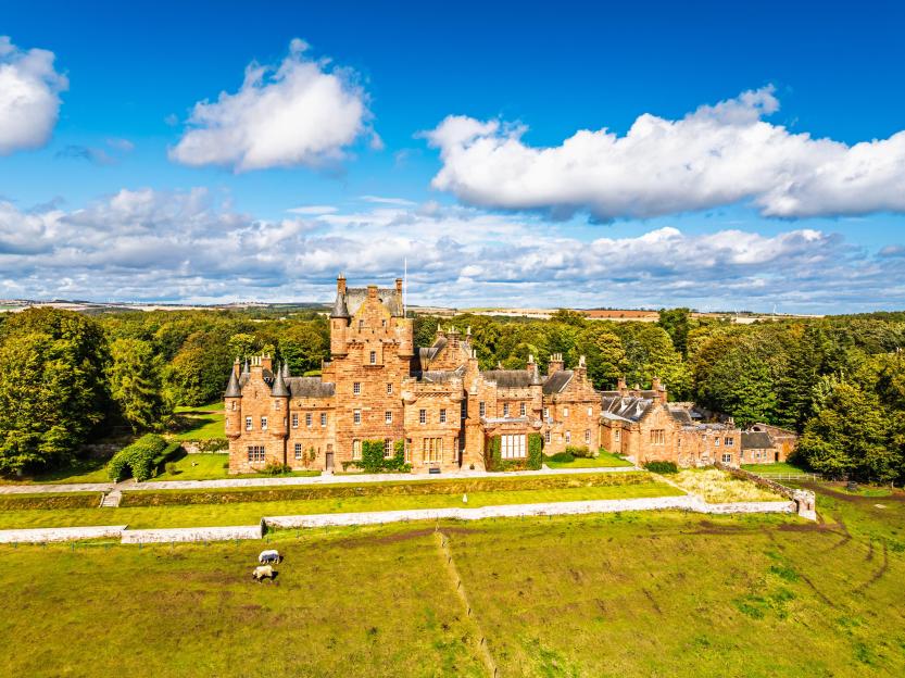 Ayton Castle, a historic brick structure with multiple towers and turrets, surrounded by lush green fields and trees under a partly cloudy sky.