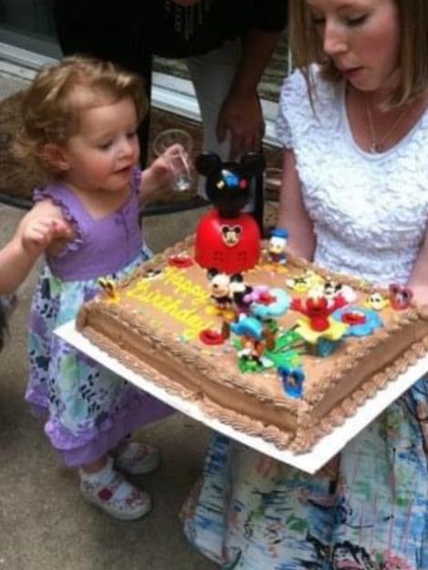 A young child with curly hair in a purple dress reaches for a Mickey Mouse birthday cake being held by a woman.