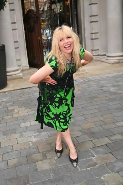 Paloma Faith smiling, wearing a black and green dress with an ornate necklace and black shoes with silver studded toes.