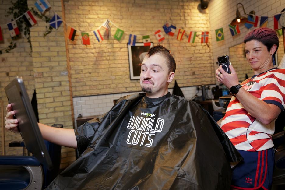 A man with a mohawk in a barber's chair looks in a mirror held by a woman with a phone.
