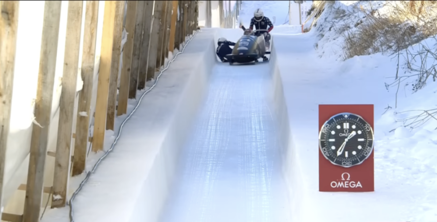 Two athletes in a bobsled on a snowy track with an Omega clock by the side.