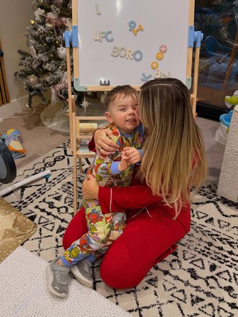 A woman in red pajamas kneeling on the floor, embracing a young boy in colorful pajamas, with a Christmas tree and whiteboard in the background.