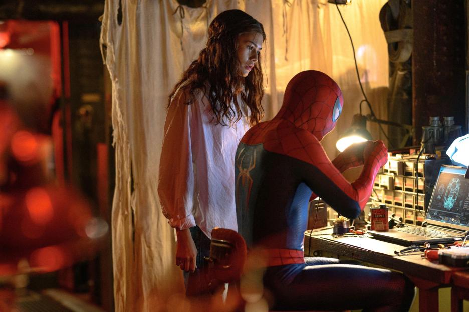 Spider-Man (Tom Holland) works on a device at a desk as Zendaya watches.