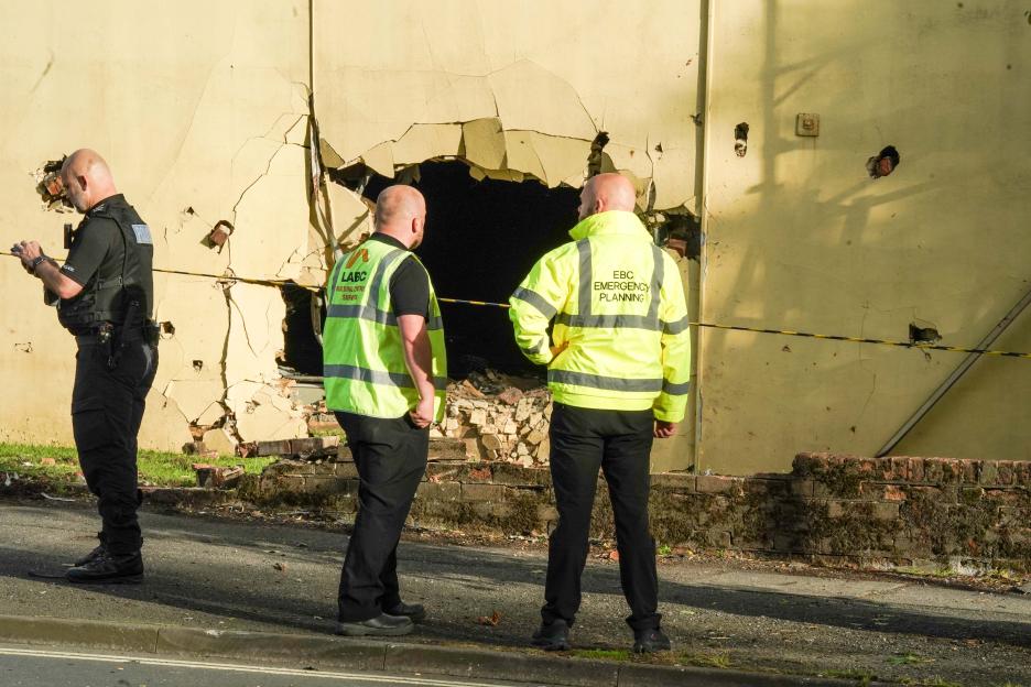 Three men, including a police officer, standing outside a building with a large hole in its wall from a car crash.