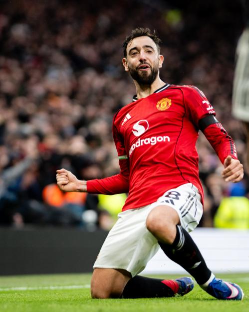 A soccer player in a red jersey and white shorts celebrates on his knees with a fist pump.