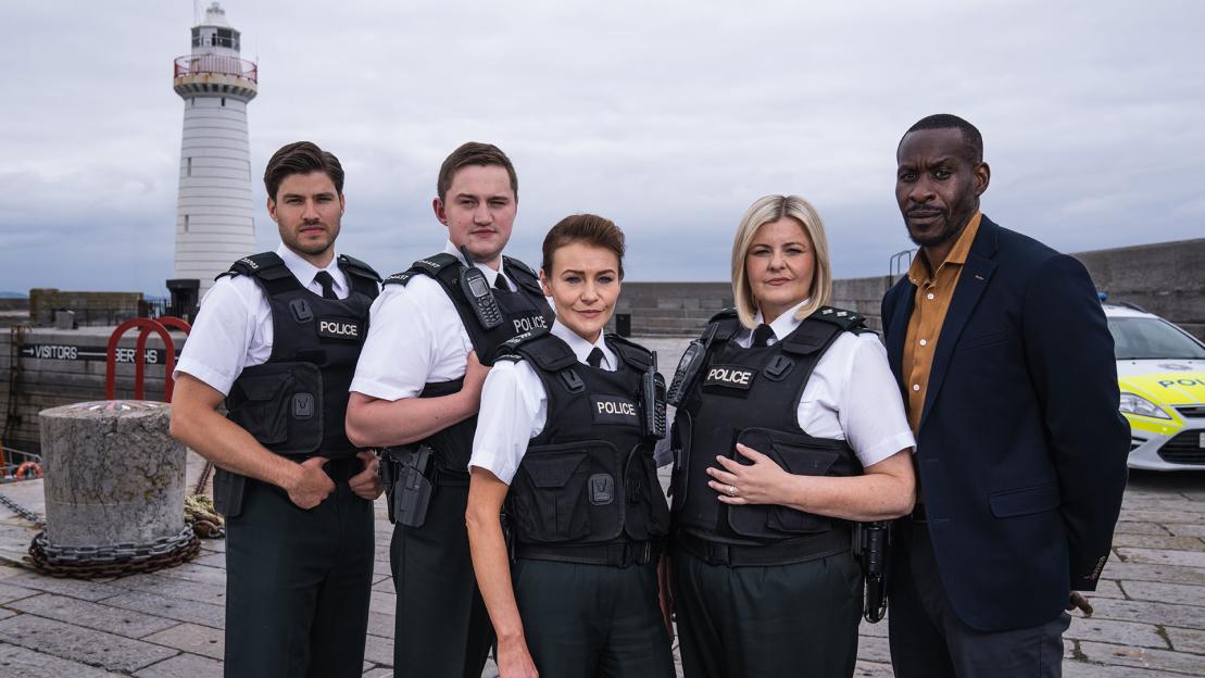Four police officers and a man in a suit stand by a lighthouse and a patrol car.